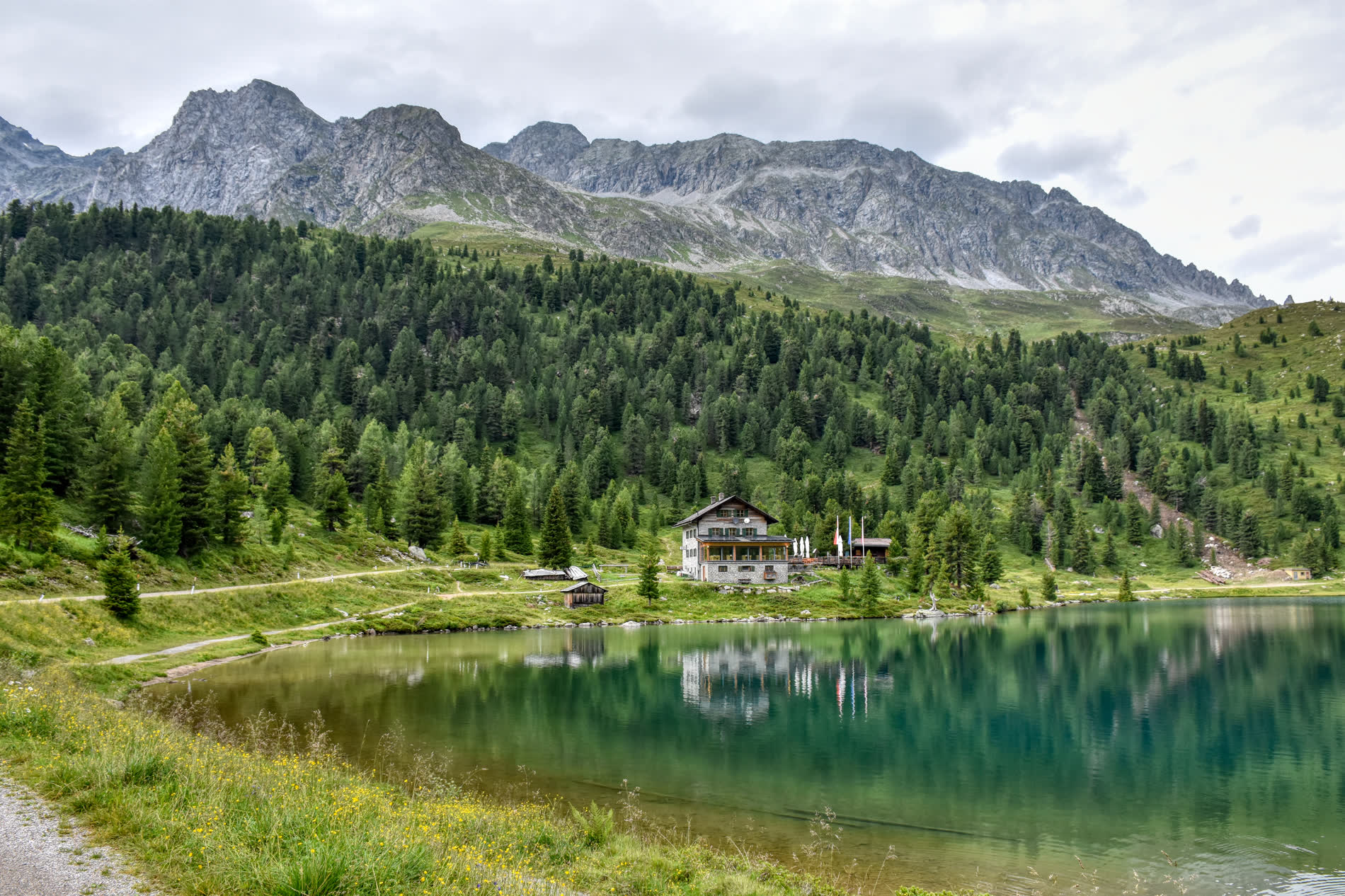 Grüner Bergsee vor Fichtenwald, am Ufer ein Gasthaus, dahinter ein felsiger Grat